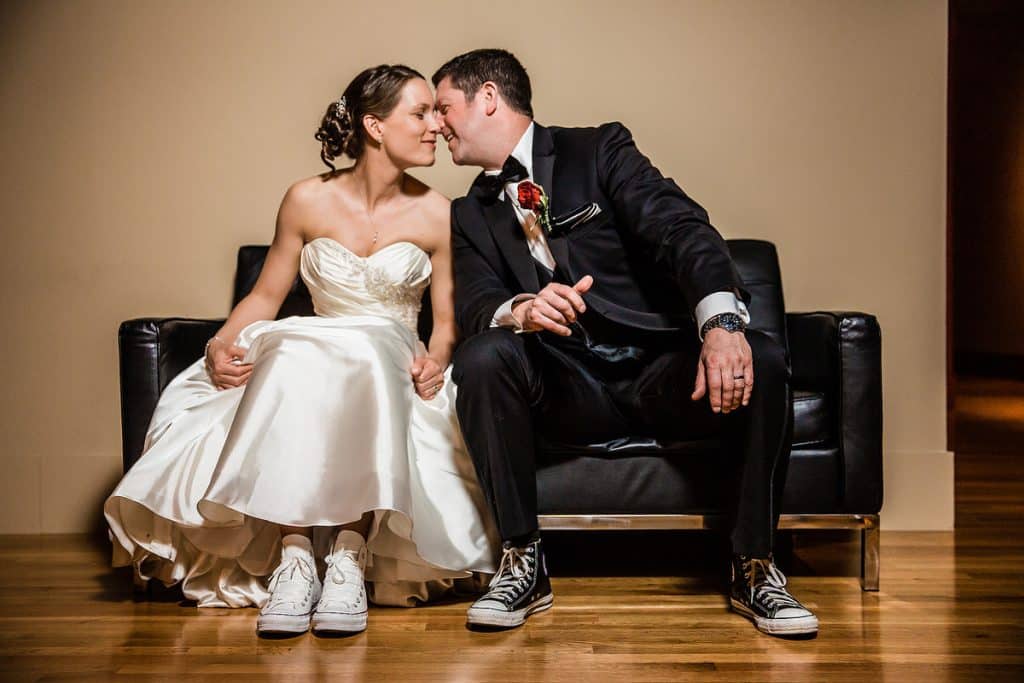 Bride and groom sitting together on a black leather couch laughing, groom wearing colorful mismatched sneakers