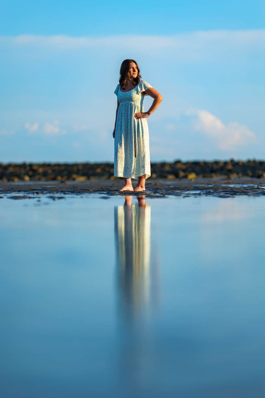 Senior portrait of a young woman in a flowing dress standing on a beach under a pastel sky
