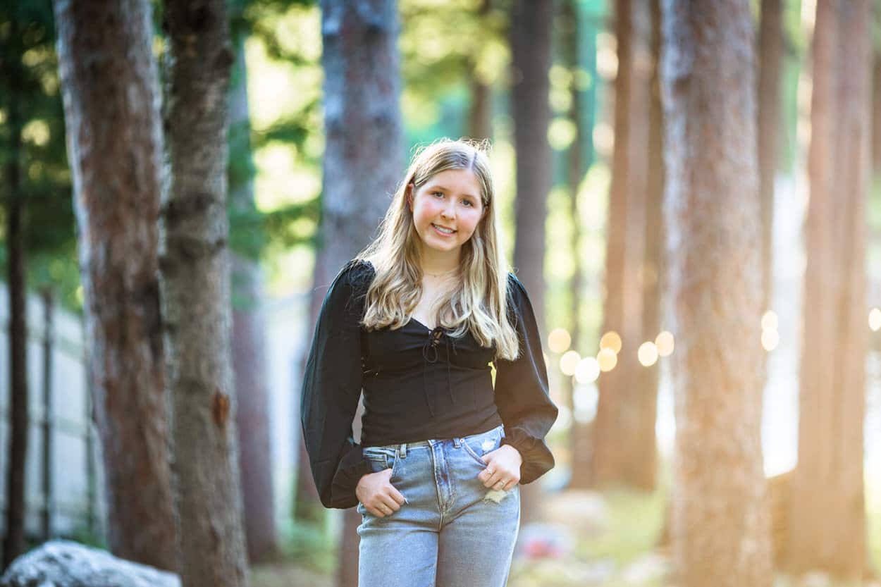Senior portrait of a young woman in a black top standing among evergreen trees with sunlight streaming through
