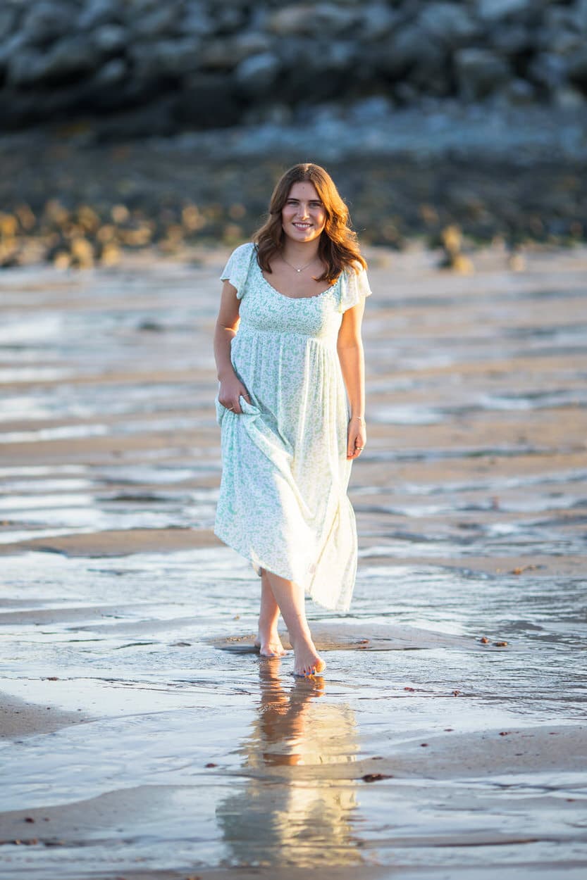 Senior portrait of a young woman in a white dress walking along a beach at low tide