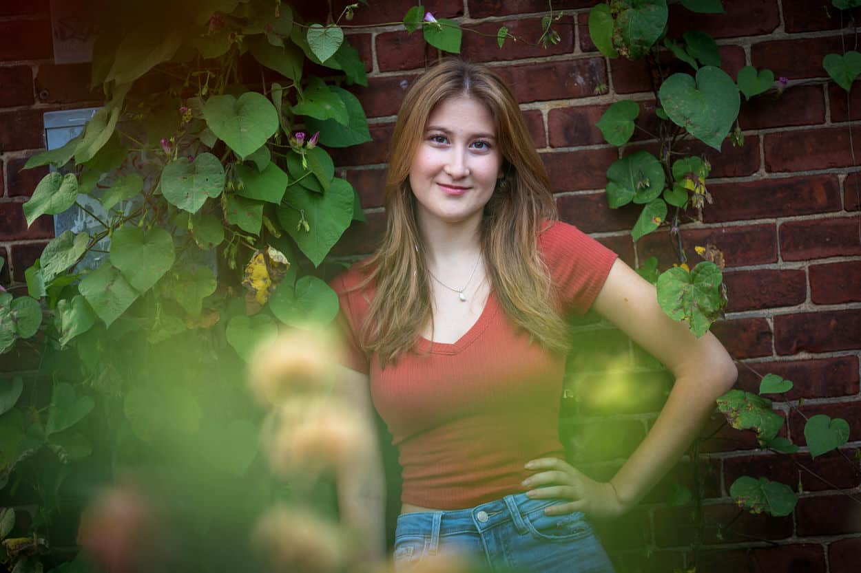 Senior portrait of a young woman in a red top standing among an ivy-covered brick wall