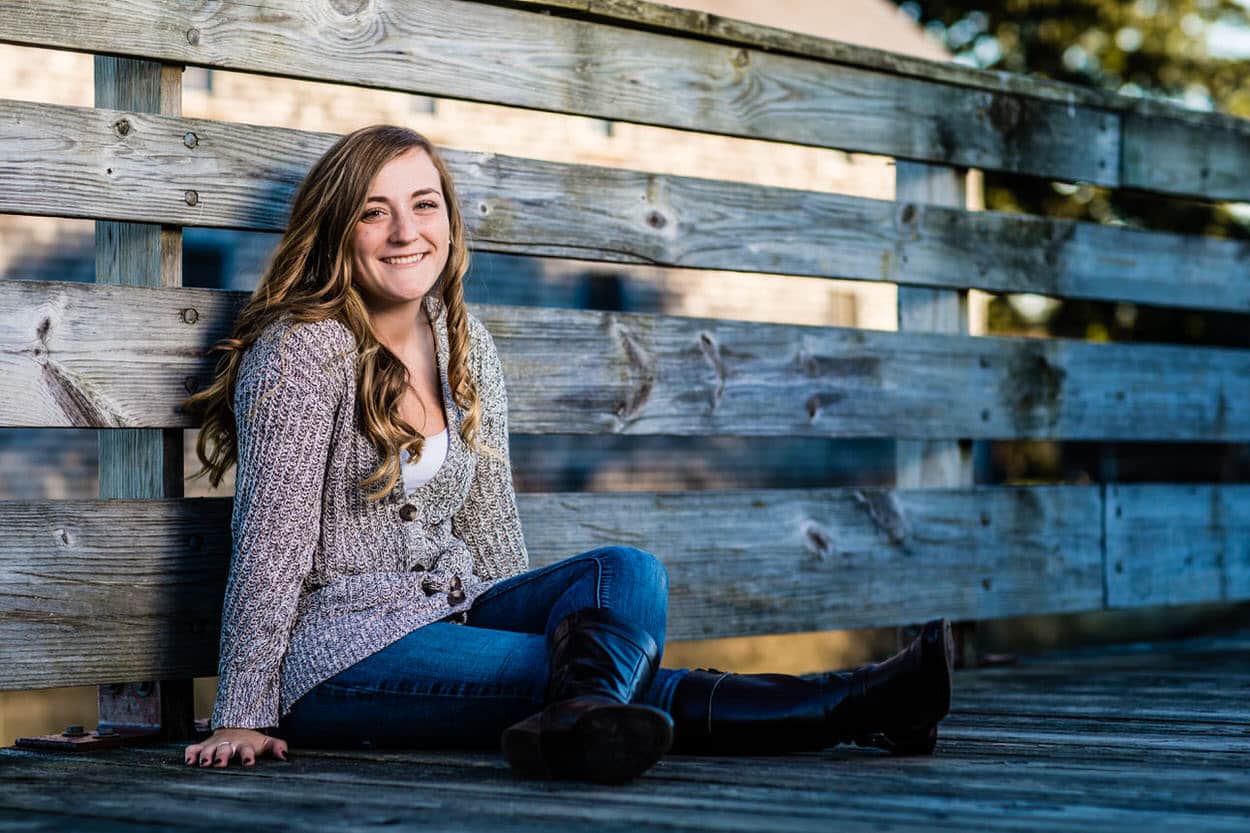 Senior portrait of a young woman sitting on a wooden fence, wearing a floral top and jeans