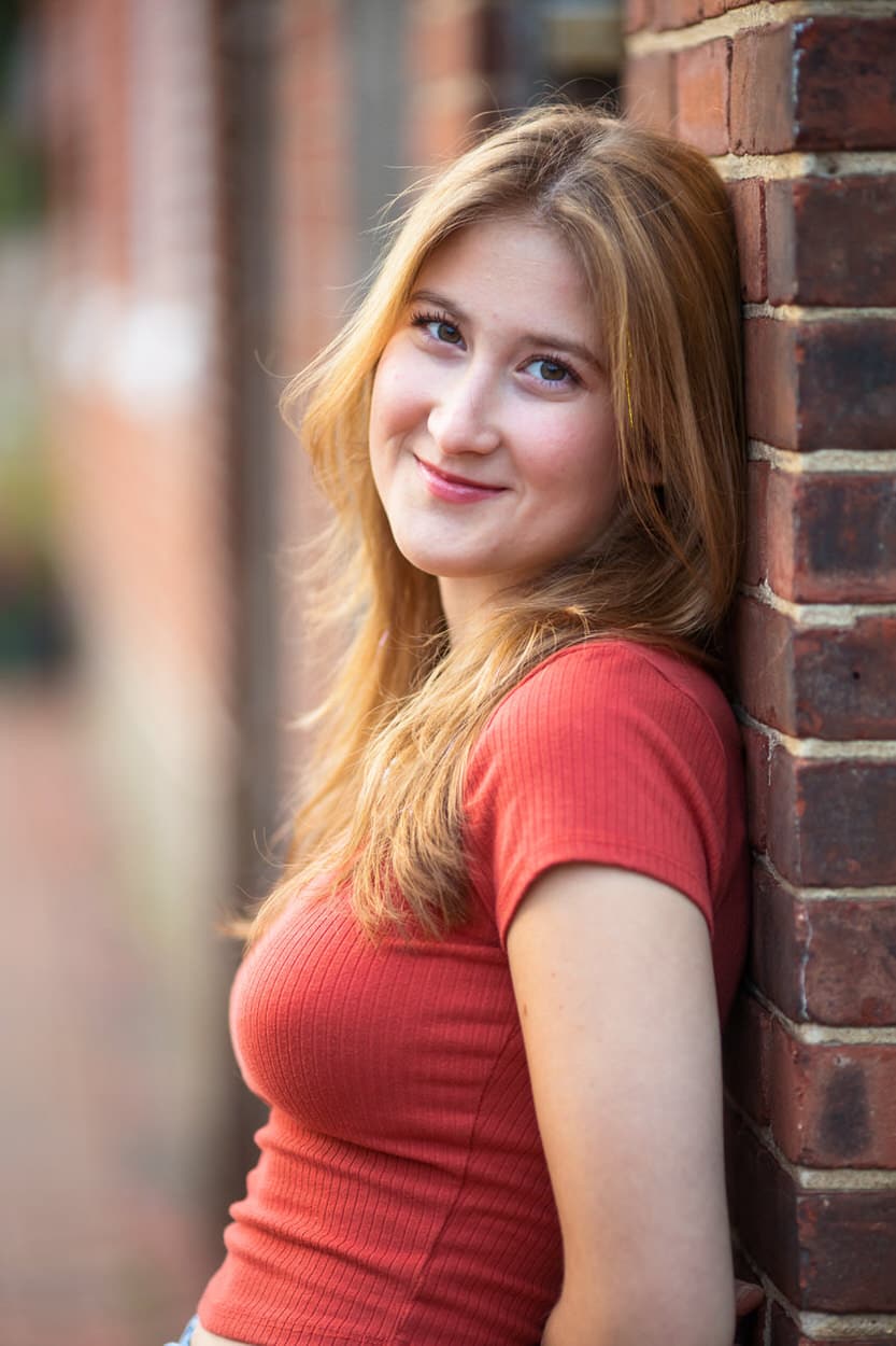 Close-up senior portrait of a young woman leaning against a brick wall wearing a red top