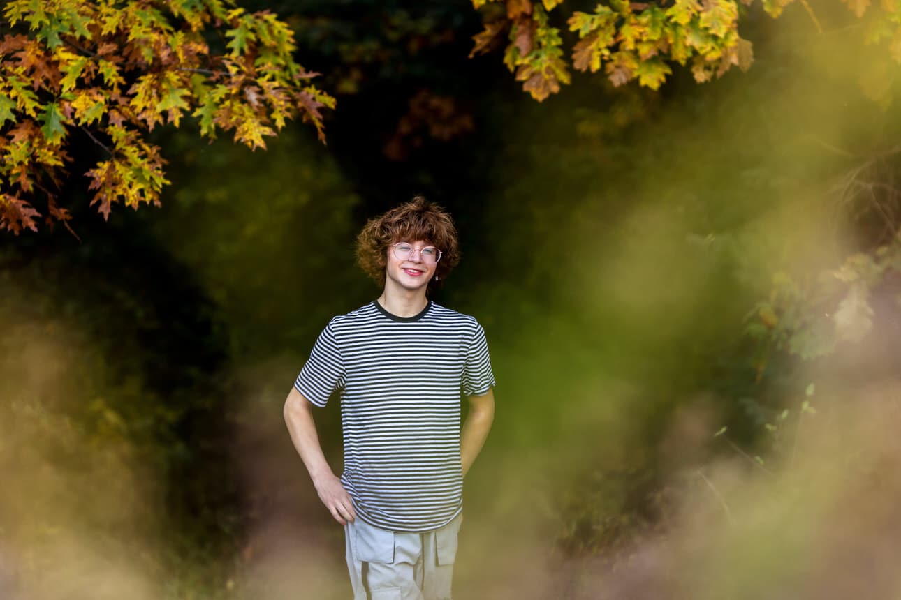 Senior portrait of a young man in a striped shirt standing among autumn foliage