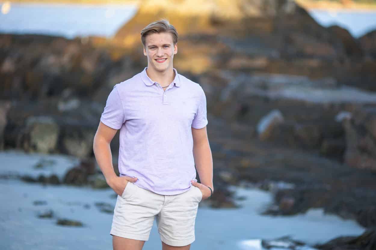 Senior portrait of a young man in a purple shirt standing on a rocky beach