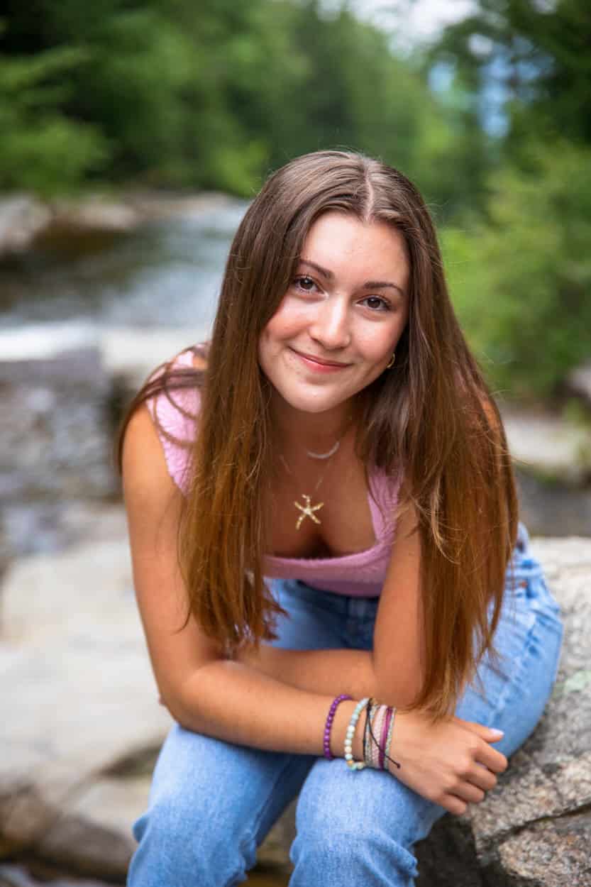 Senior portrait of a young woman sitting on rocks by a forest stream, wearing a pink top