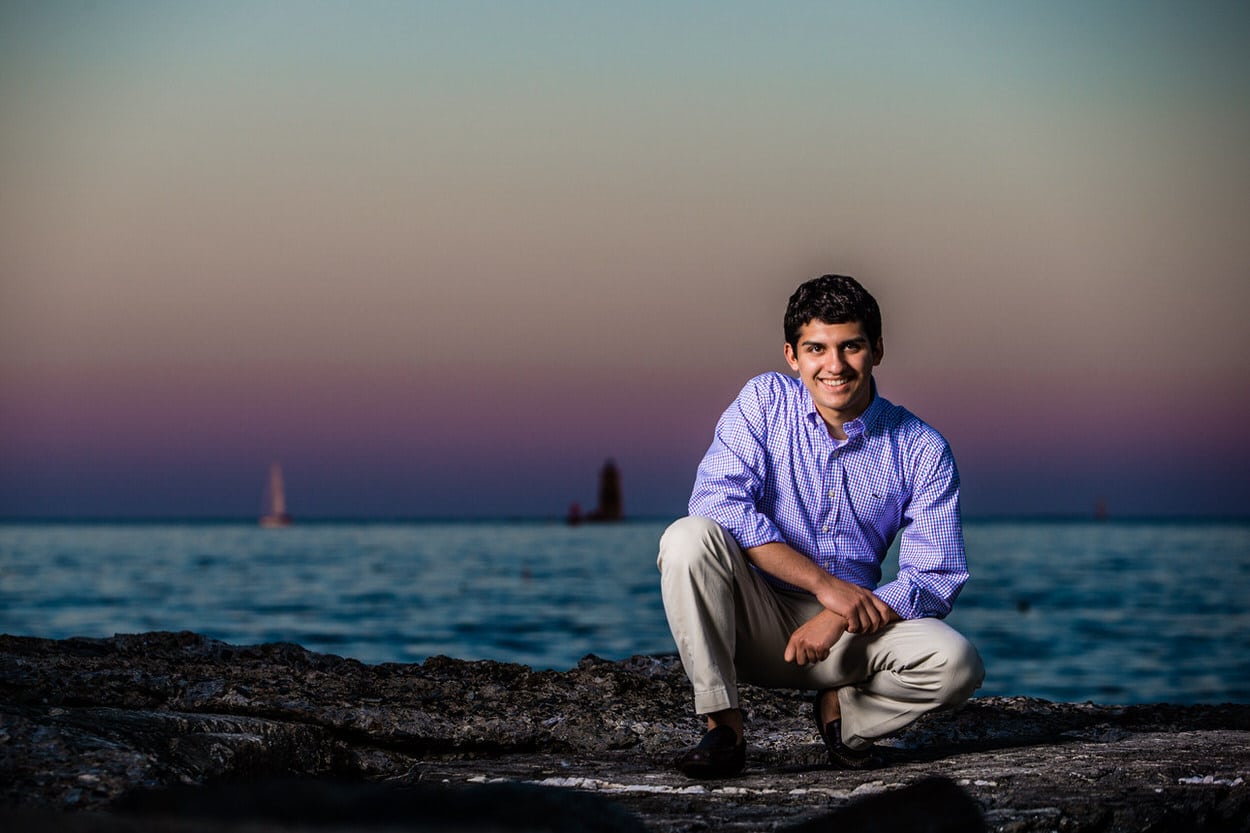 Senior portrait of a young man sitting on a rock by the water at sunset