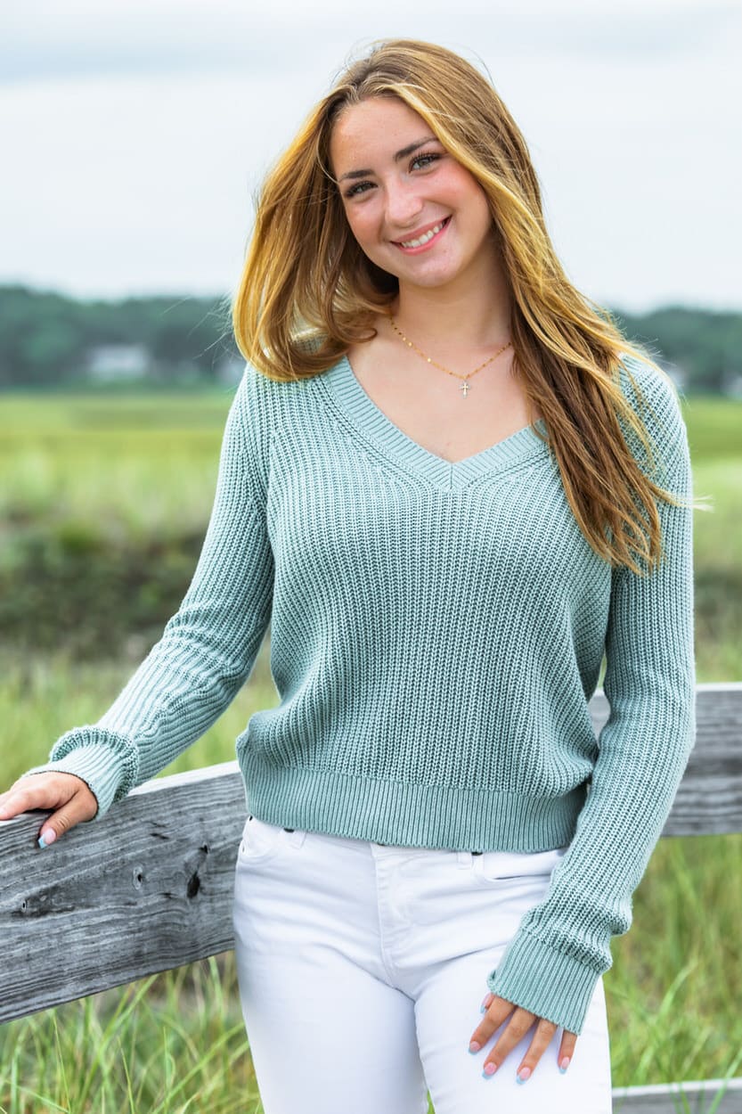 Senior portrait of a young woman in a sage green sweater leaning against a wooden fence
