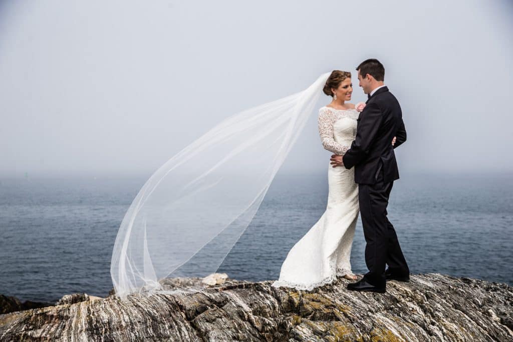 Bride and groom embracing on a rocky coastal cliff, bride's long veil blowing in the wind against a foggy ocean backdrop
