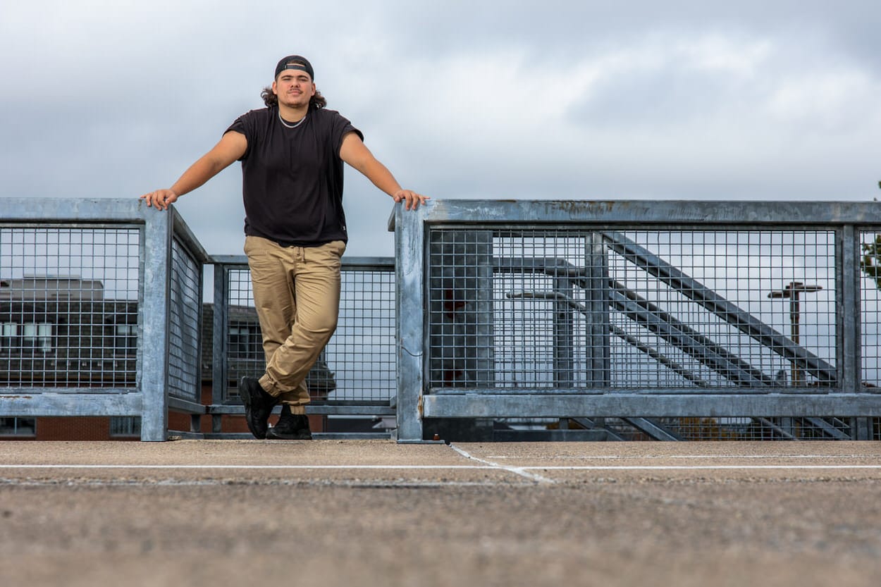 Senior portrait of a young man in a black jacket standing on a metal walkway bridge at dusk
