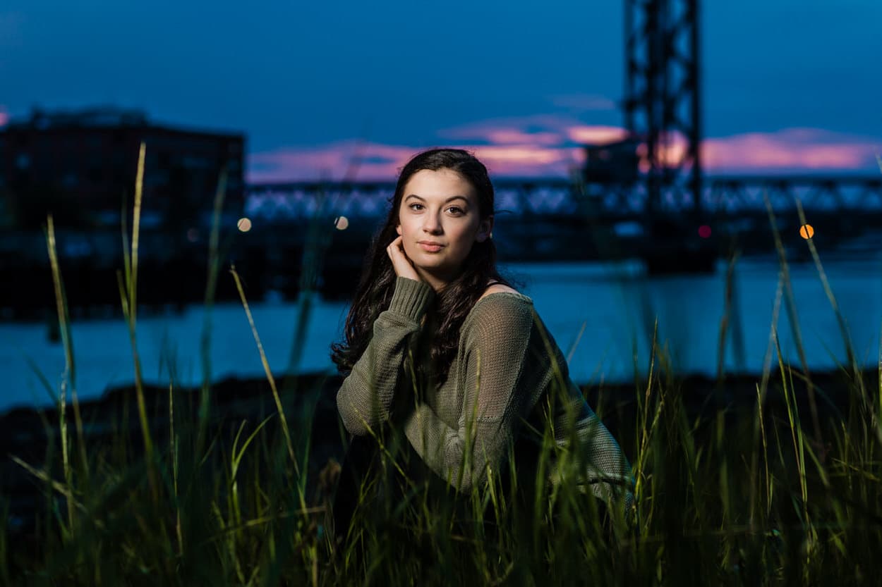 Senior portrait of a young woman sitting among reeds at dusk with a bridge in the background
