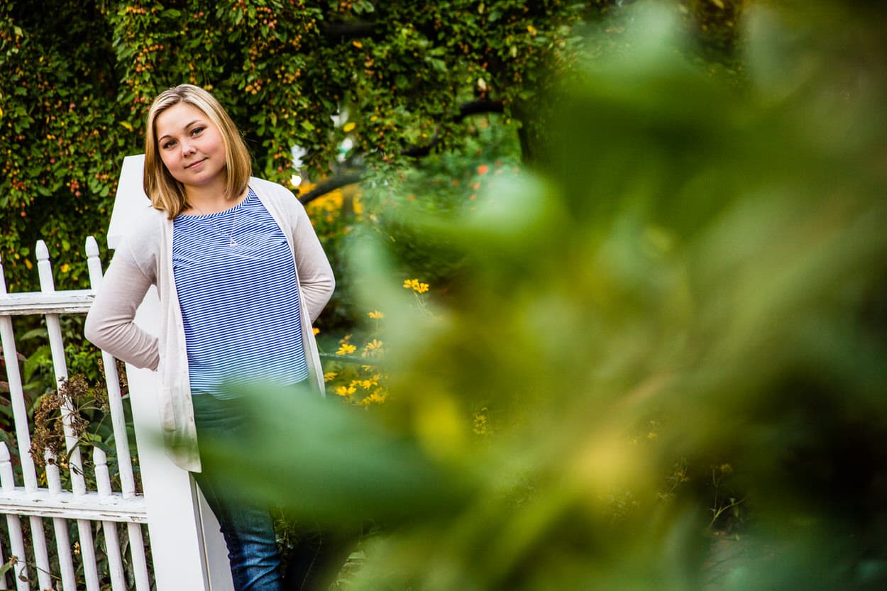 Senior portrait of a young woman in a white cardigan standing by a white picket gate, framed by green foliage
