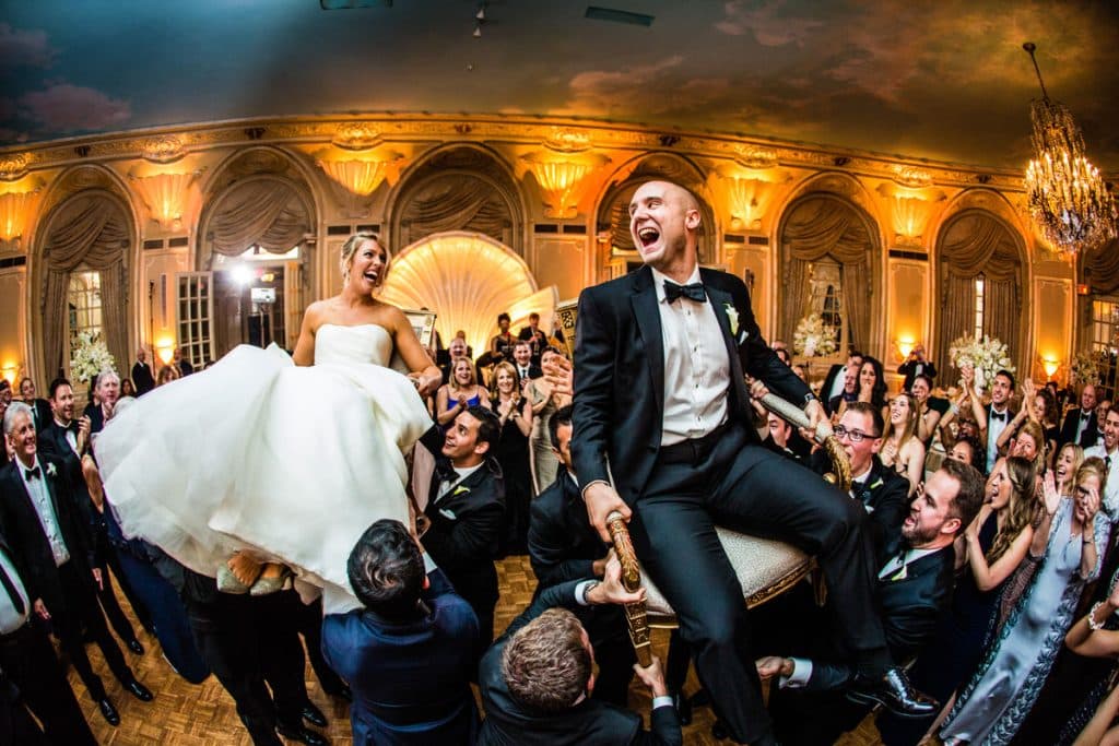 Groomsmen lifting the groom into the air to celebrate at an elegant reception venue strung with lights
