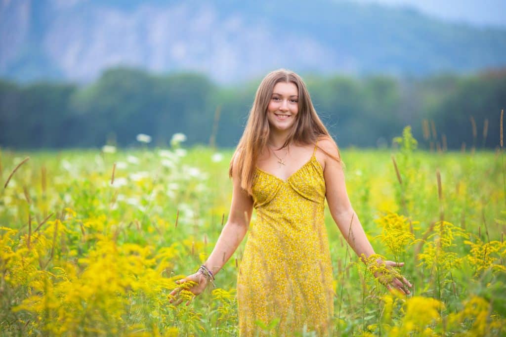 Senior portrait of a young woman in a yellow dress standing in a wildflower field with mountains behind her