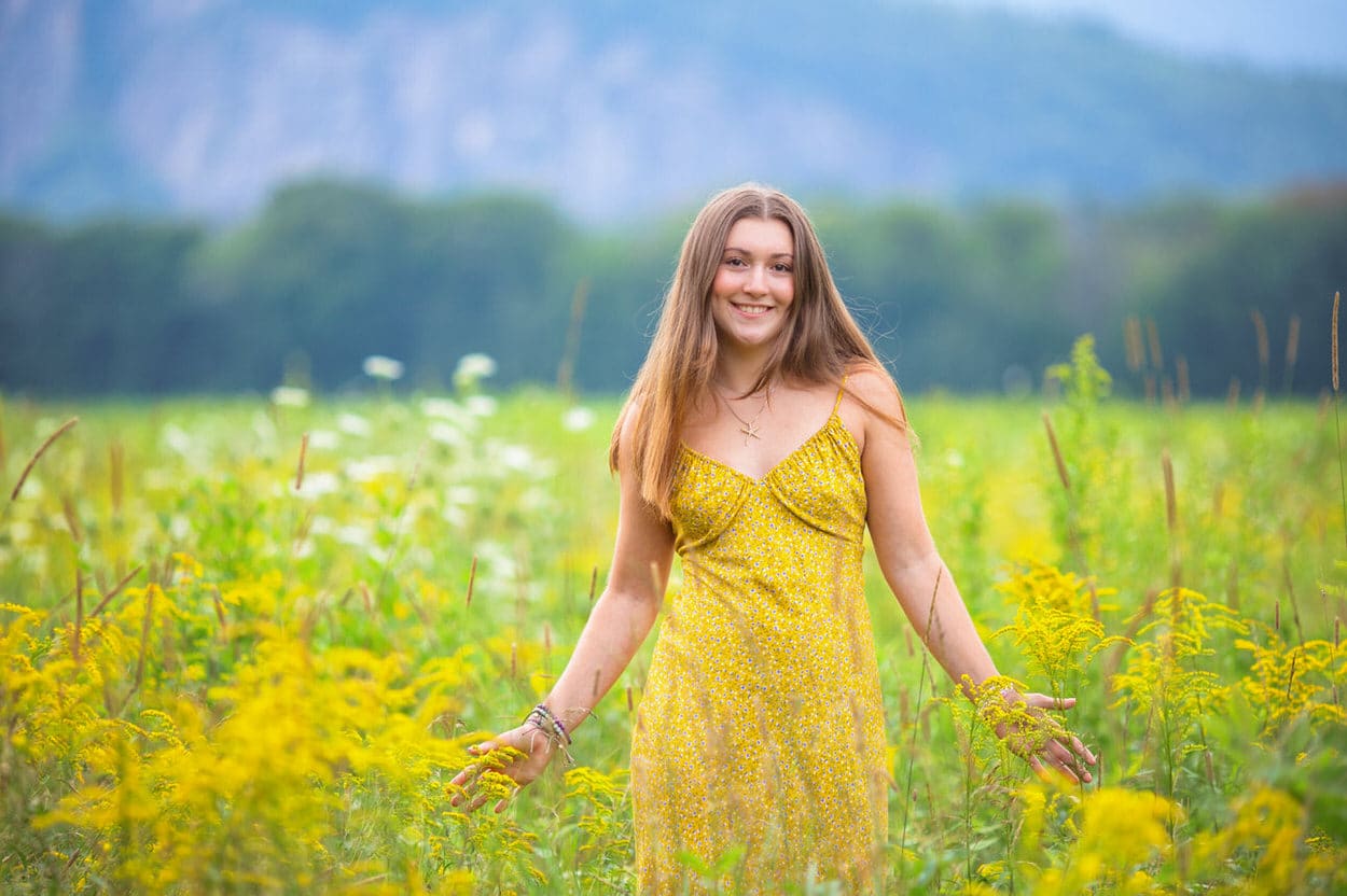 Senior portrait of a young woman in a yellow dress standing in a wildflower field with mountains behind her