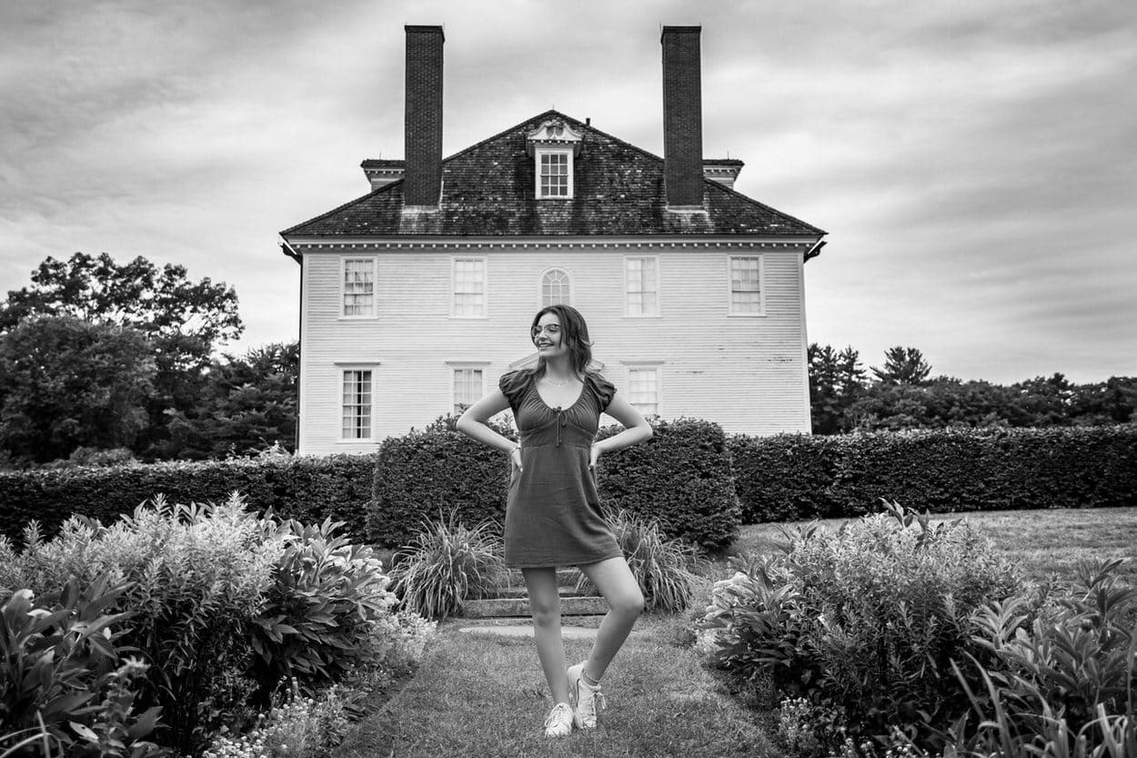 Black and white senior portrait of a young woman in a flowing dress standing in front of a colonial-style house
