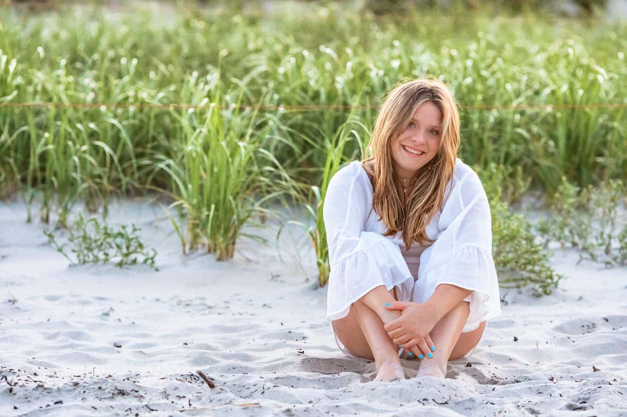 Senior portrait of a young woman in a white outfit sitting in beach grass