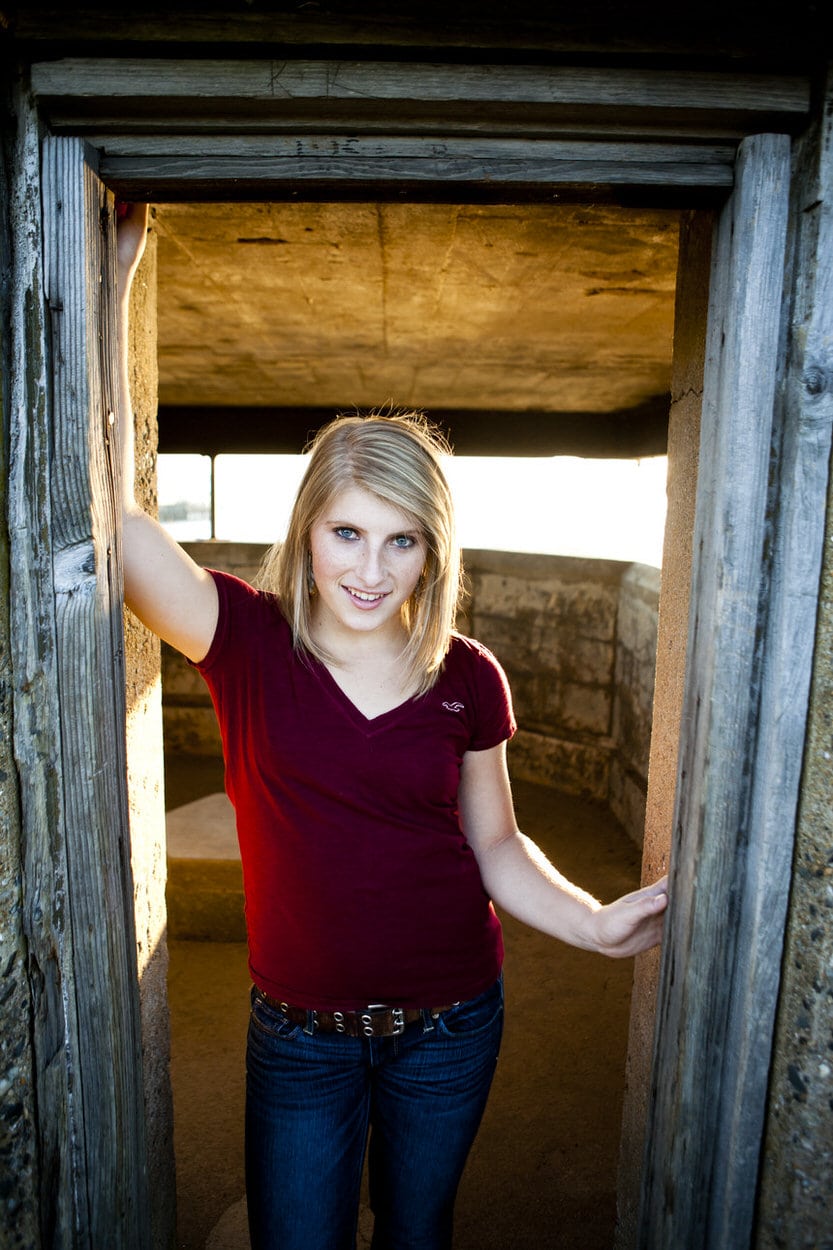 Senior portrait of a young woman in a maroon top standing in a rustic wooden barn doorway