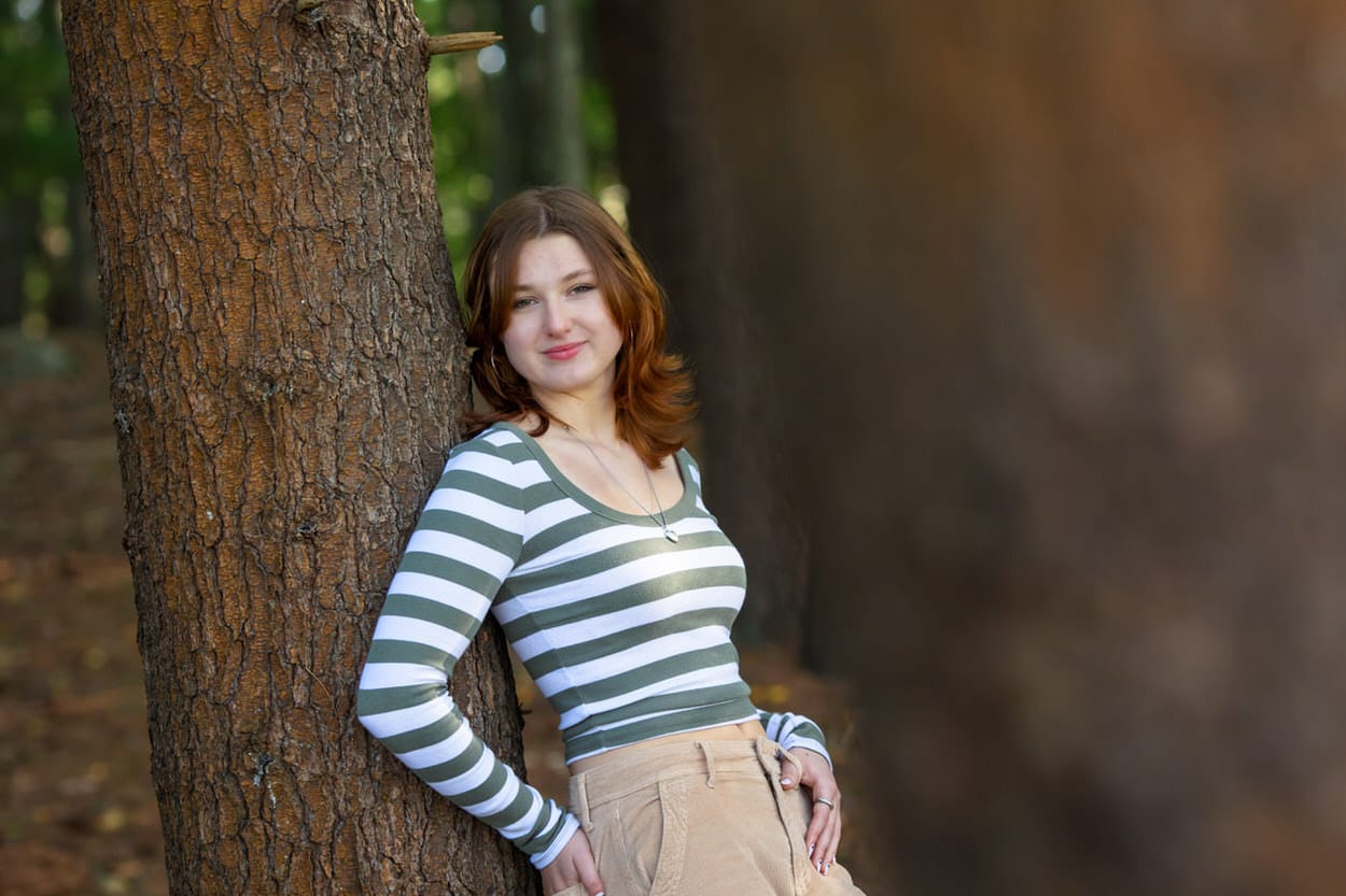 Senior portrait of a young woman in a striped sweater leaning against a tree trunk