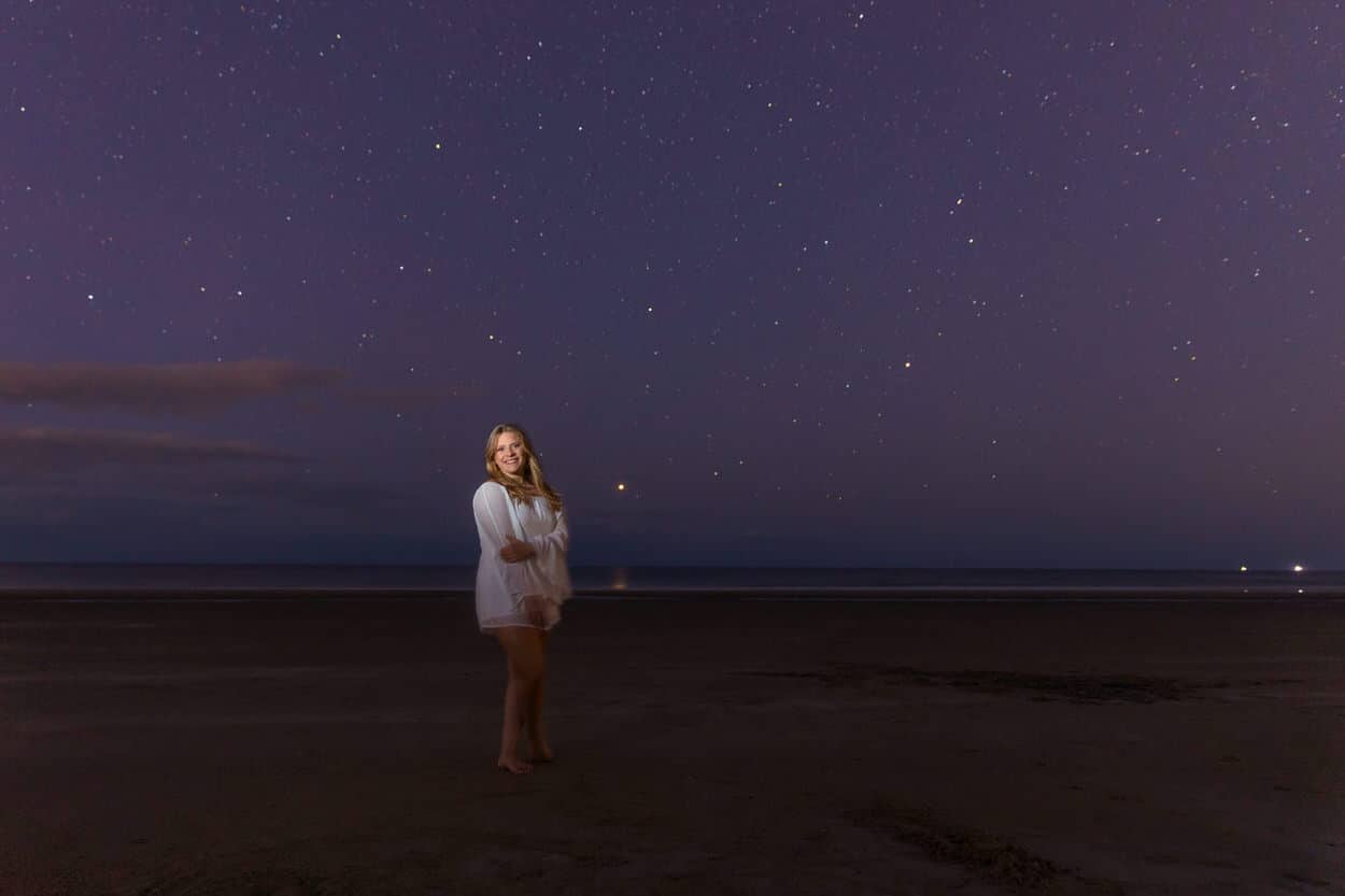 Senior portrait of a young woman in a white dress standing on a beach at night under a starry sky