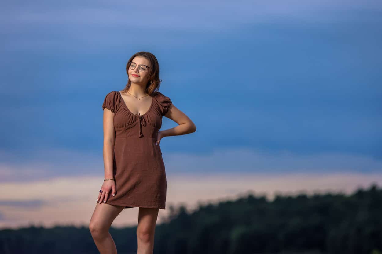 Senior portrait of a young woman in a brown dress standing outdoors at dusk near water