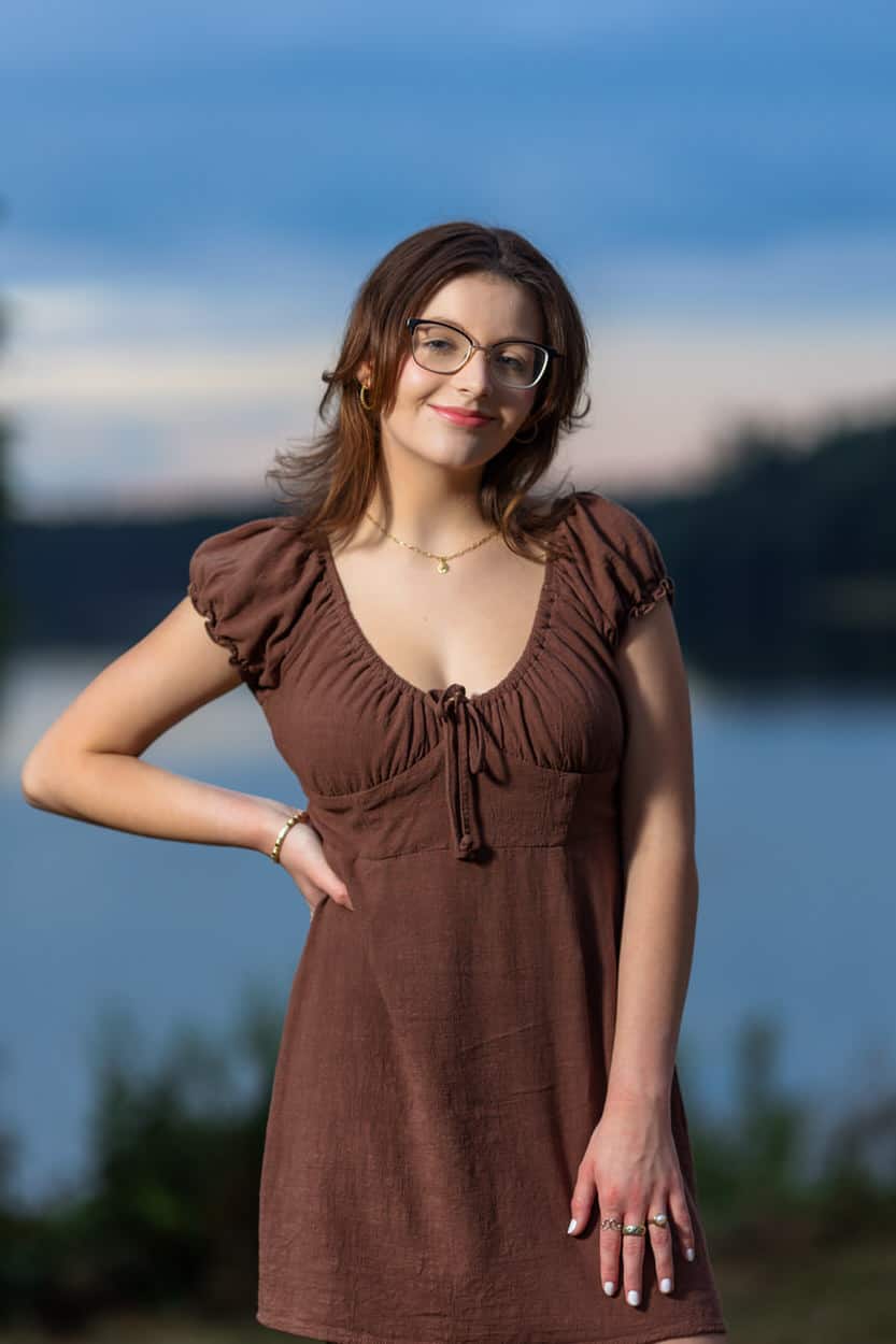 Senior portrait of a young woman wearing glasses and a brown dress, hand on hip, with water in the background