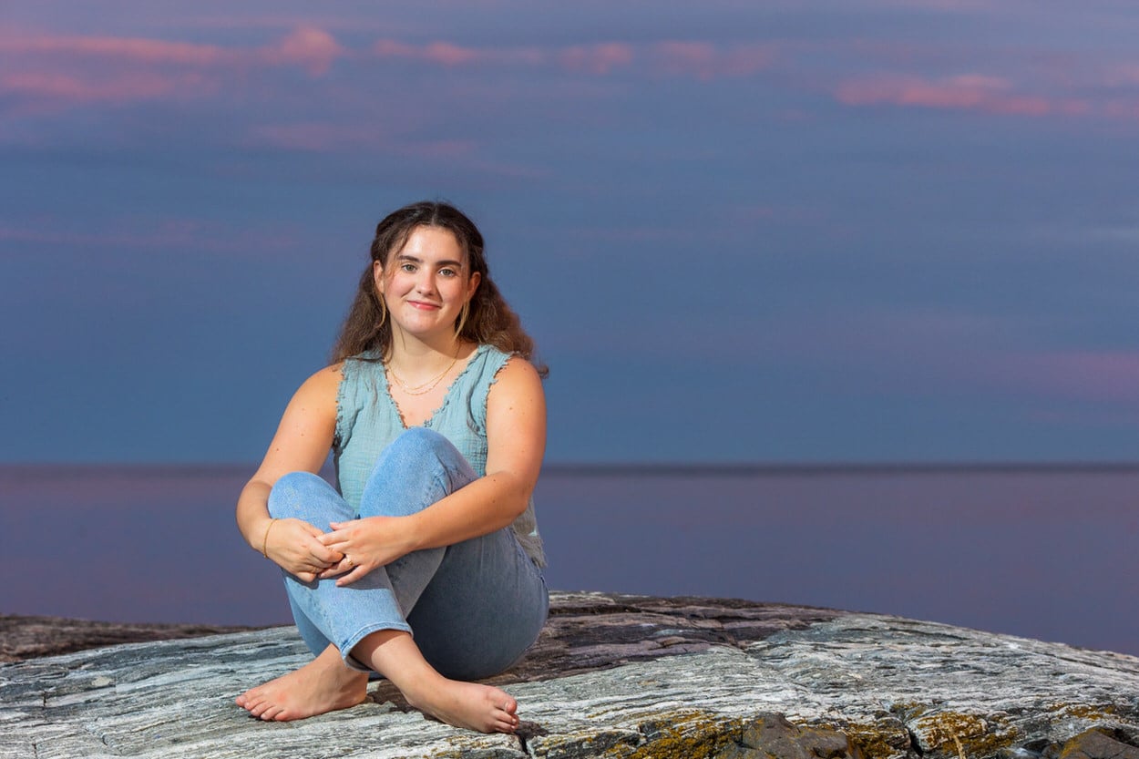 Senior portrait of a young woman sitting cross-legged on rocks at sunset by the water