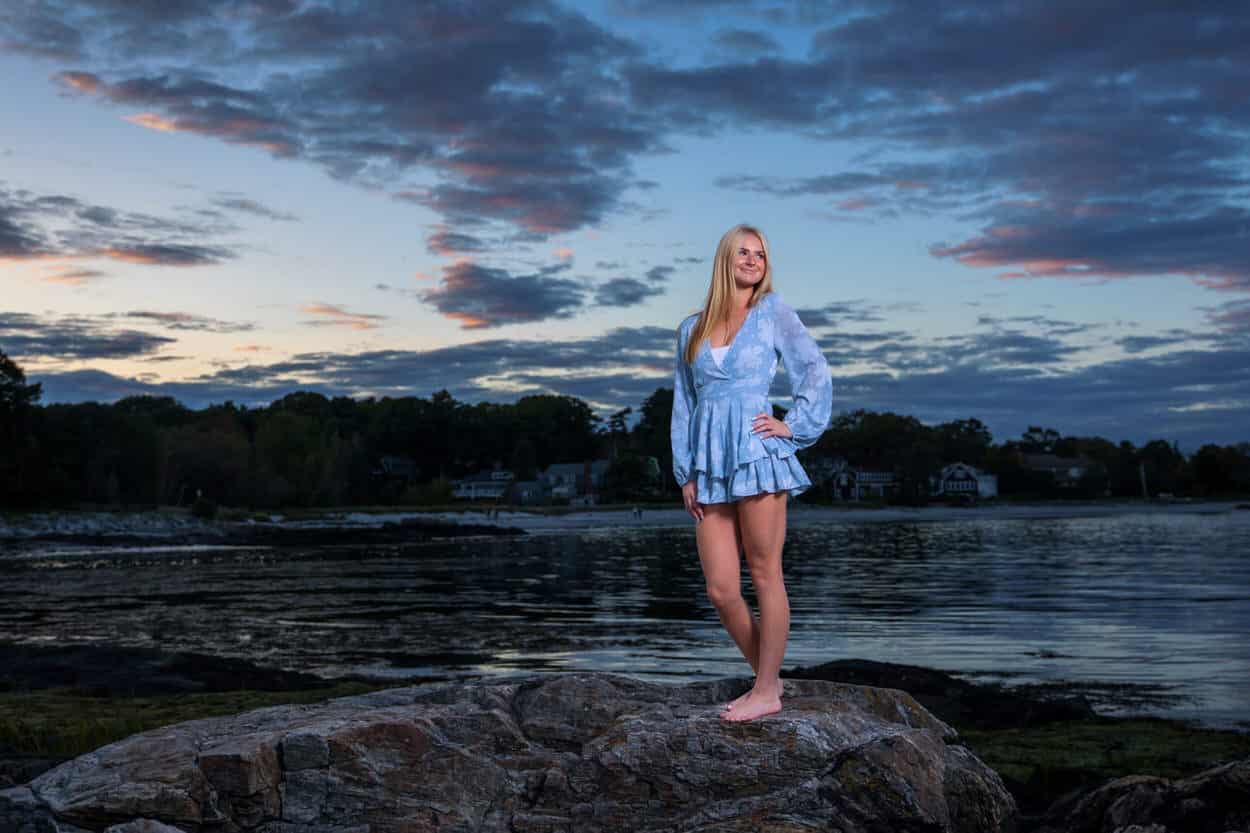 Senior portrait of a young woman standing barefoot on rocks at sunset by the water
