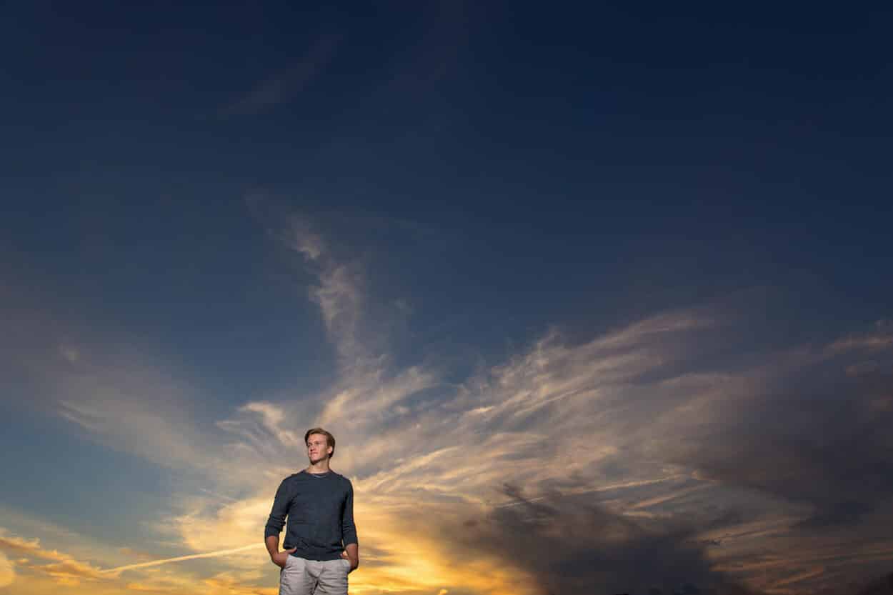 Senior portrait of a young man standing silhouetted against a dramatic sunset sky