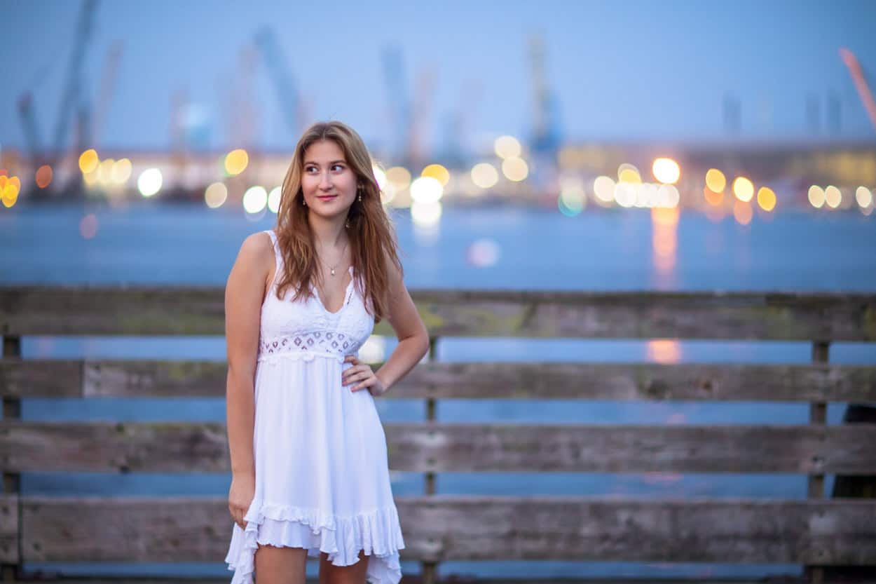 Senior portrait of a young woman standing on a wooden pier at night with distant city lights