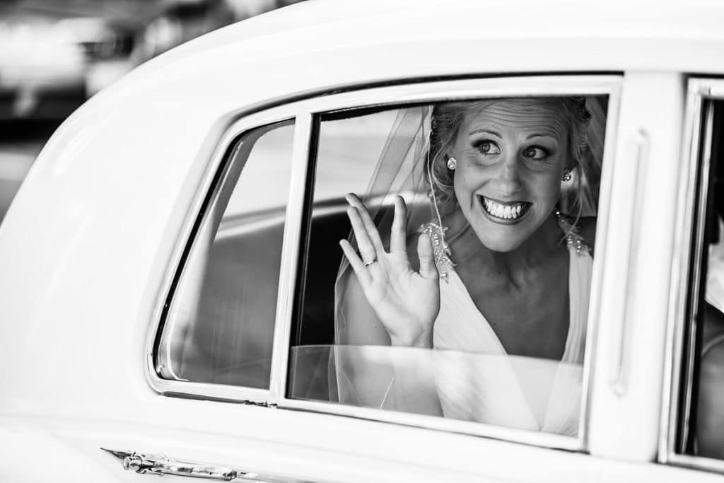 Bride waving from inside the window of a vintage white car