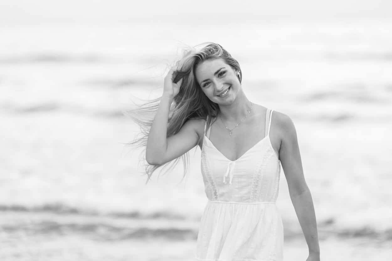 Black and white senior portrait of a young woman in a white dress with wind blowing her hair on a beach