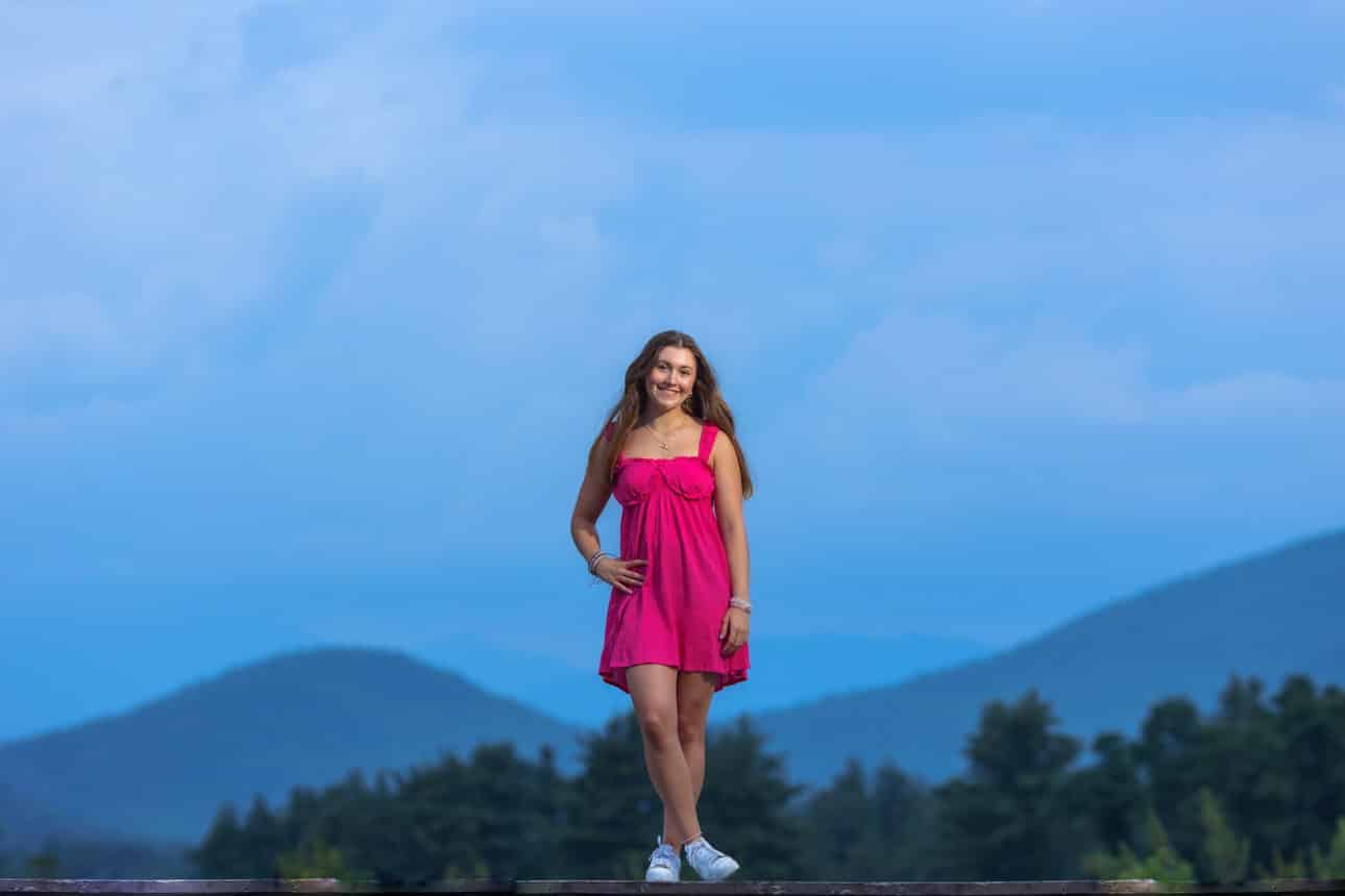 Senior portrait of a young woman in a pink dress standing on a rocky overlook with mountains behind her