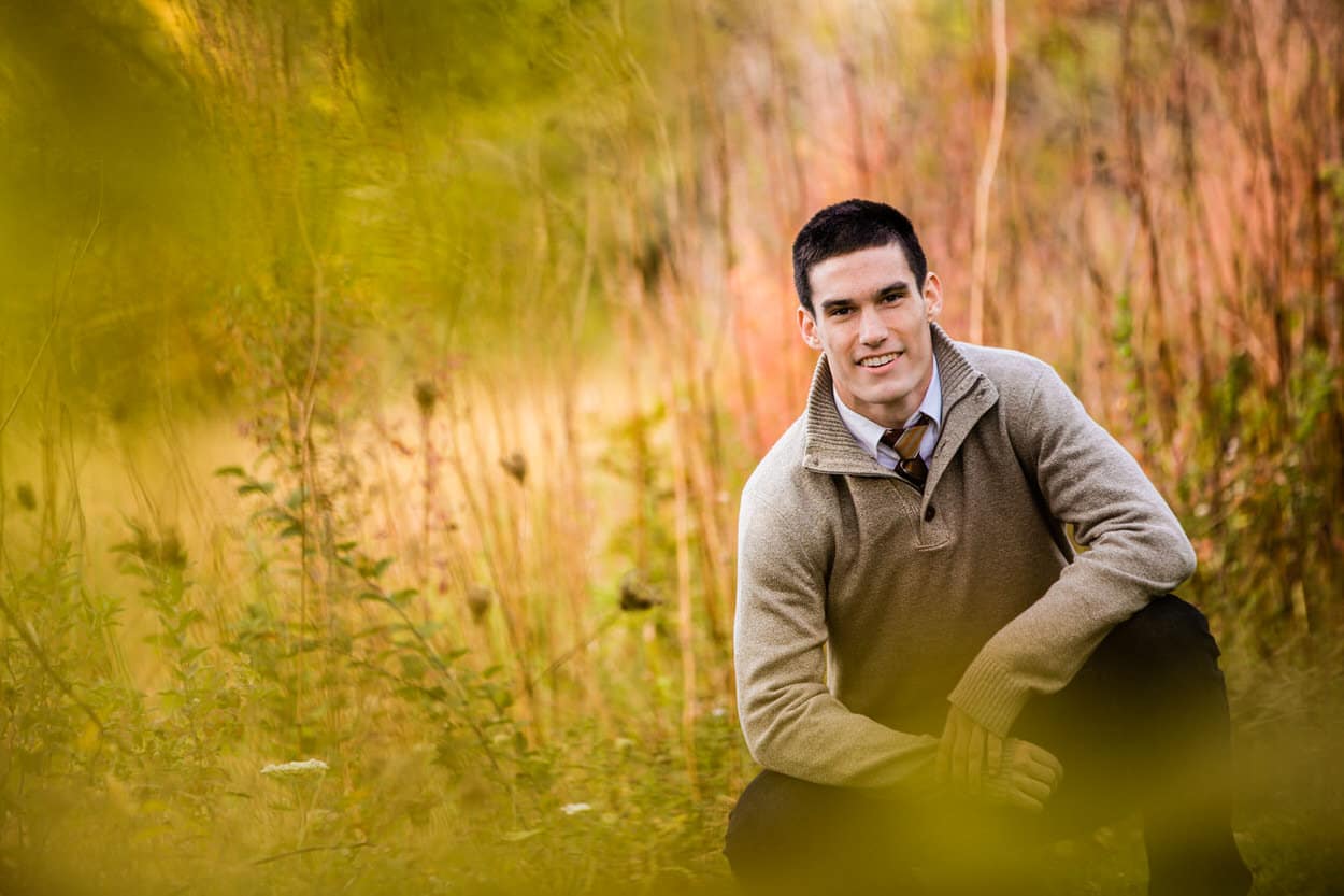Senior portrait of a young man crouching in a golden field at sunset