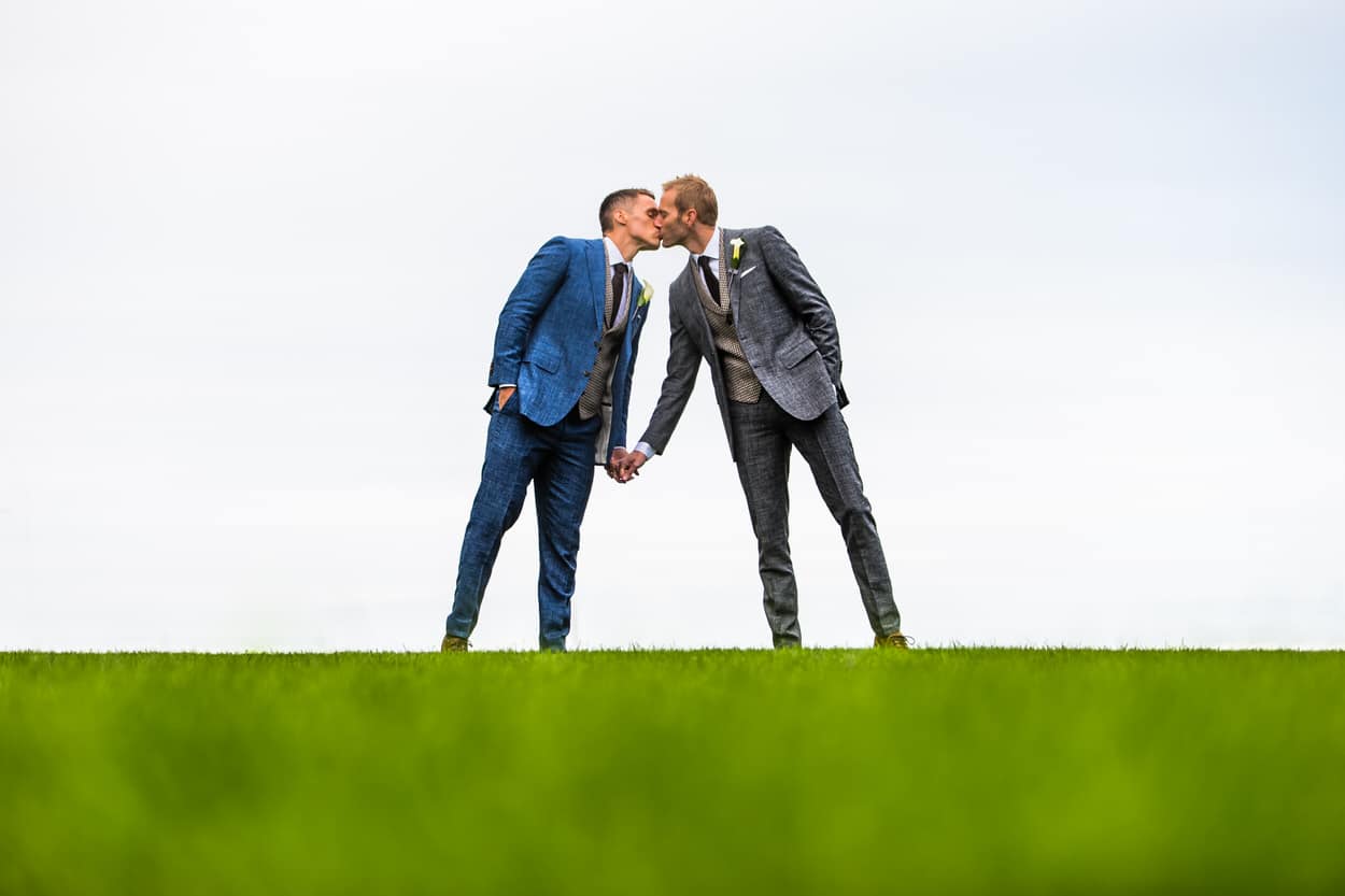 Two grooms kissing on a grassy hilltop during their elopement