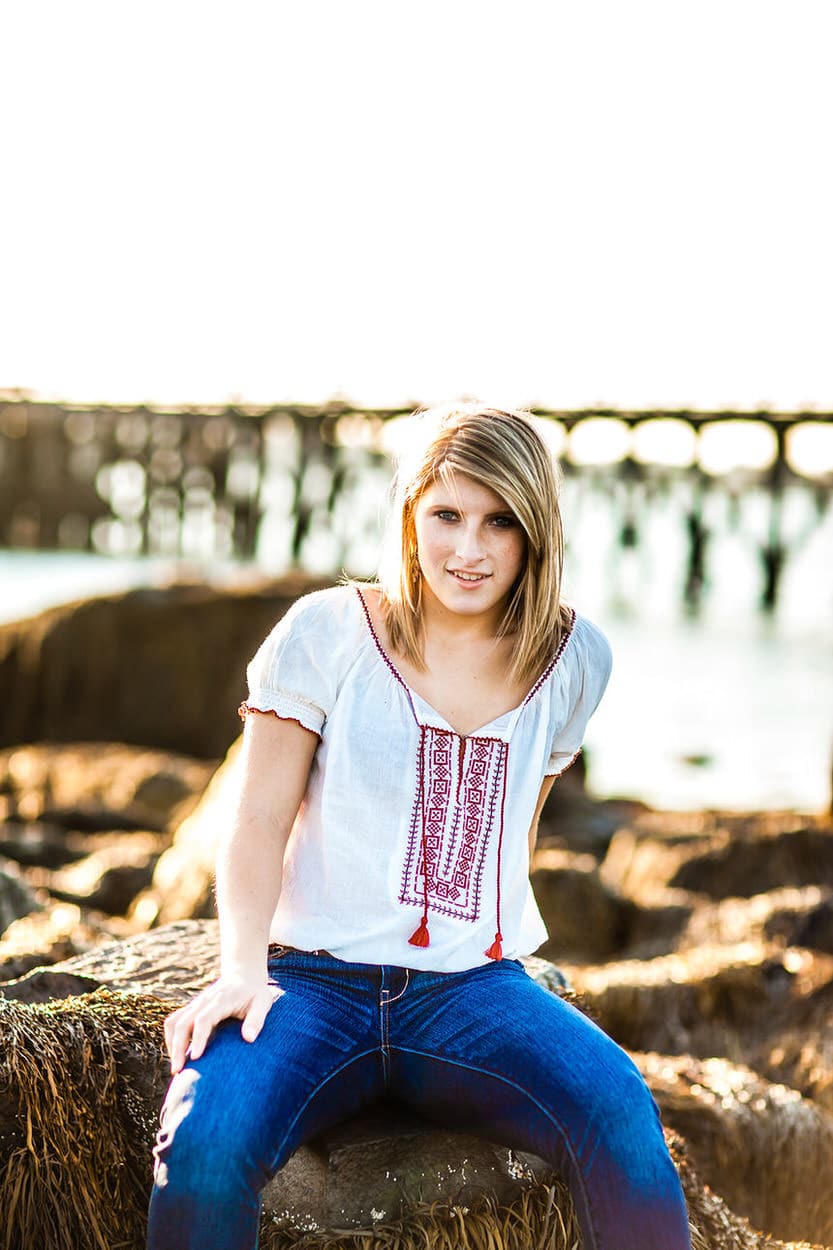Senior portrait of a young woman in a white peasant top sitting on rocks by the water