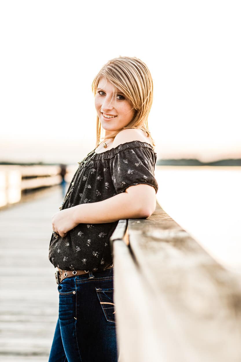 Senior portrait of a young woman leaning against a wooden railing with a body of water in the background