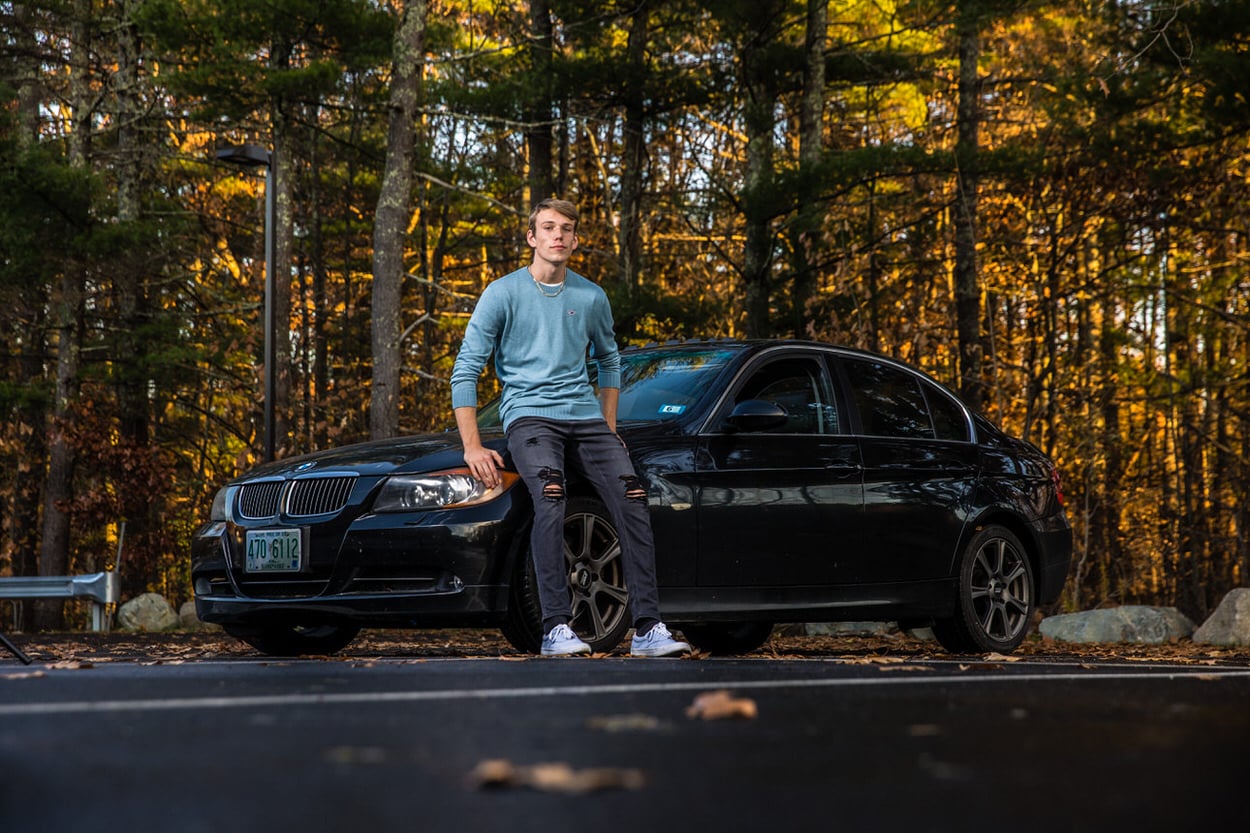 Senior portrait of a young man standing beside a black car on a forest road