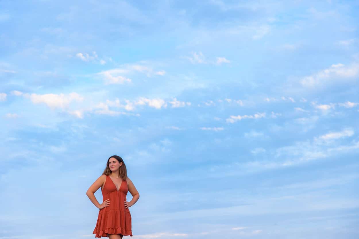 Senior portrait of a young woman in an orange dress standing on a beach under a cloudy sky