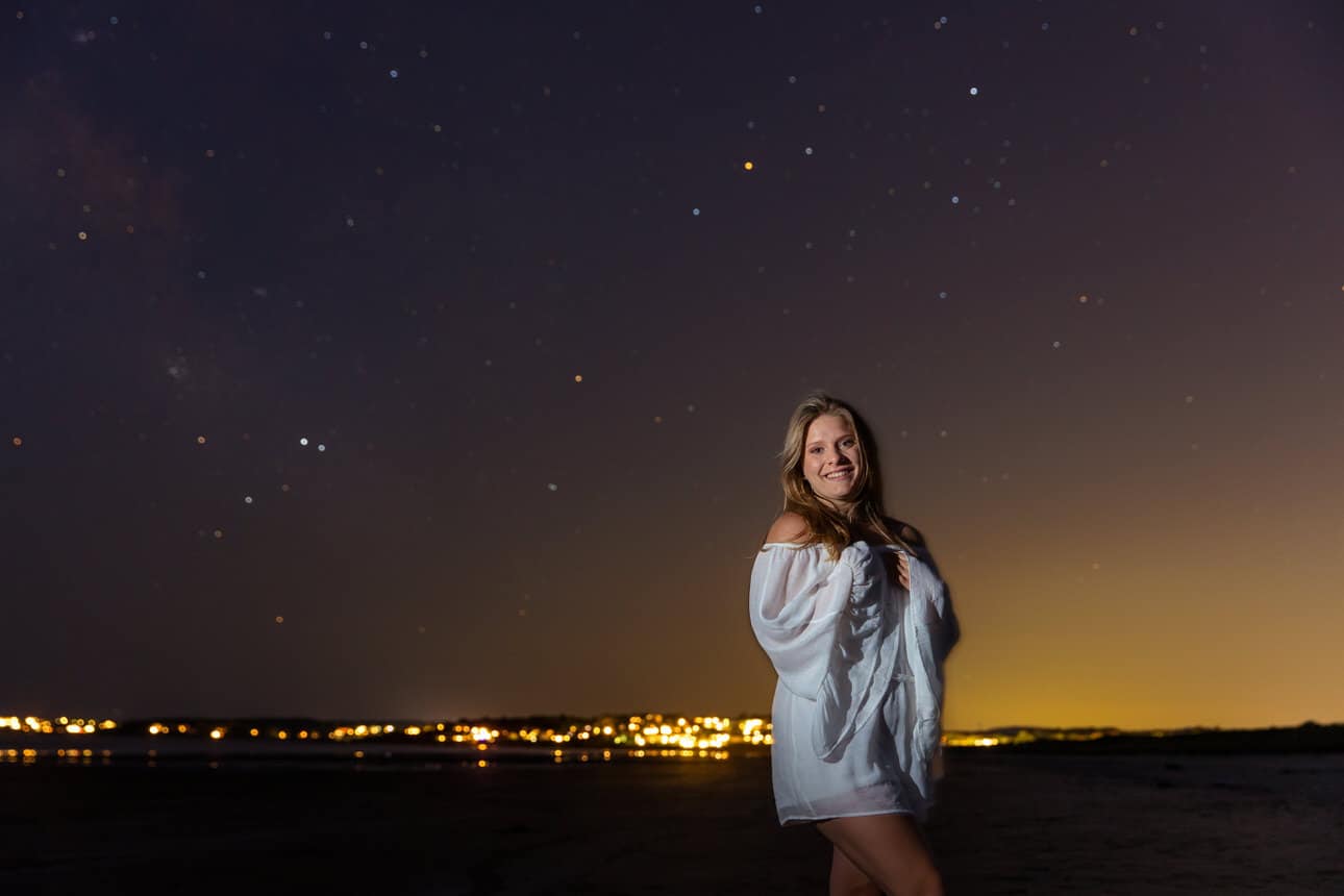 Senior portrait of a young woman standing on a hilltop at night under a starry sky with city lights below