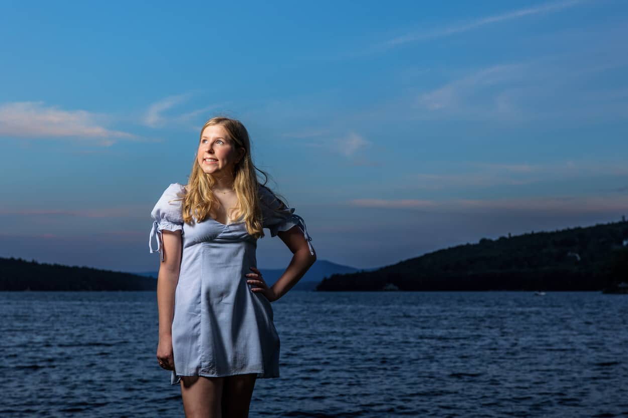 Senior portrait of a young woman in a gray dress standing on rocks by a lake, hand on hip