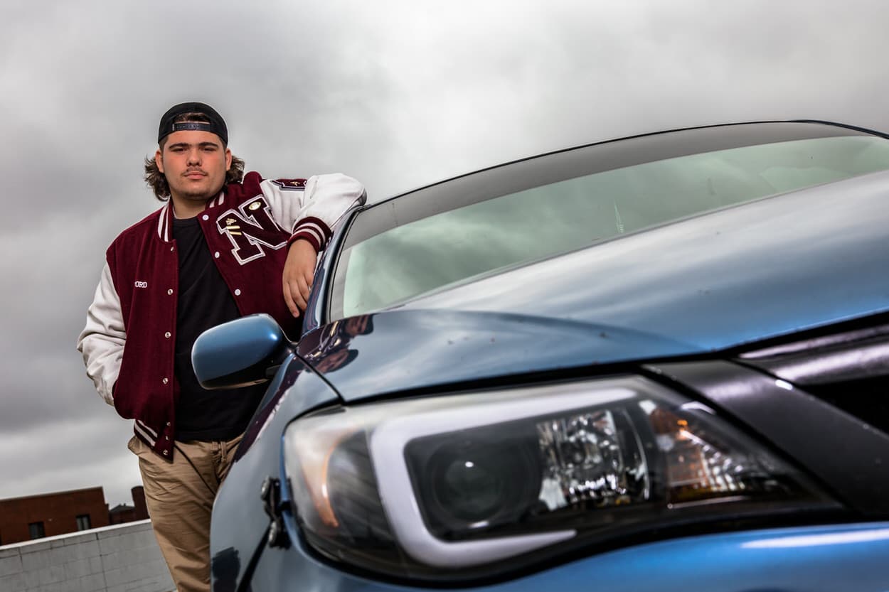 Senior portrait of a young man in a varsity jacket leaning against a car, adjusting his collar