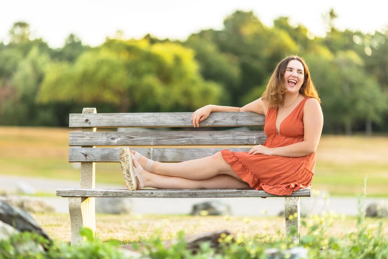 Senior portrait of a young woman in an orange dress sitting on a wooden bench in a park