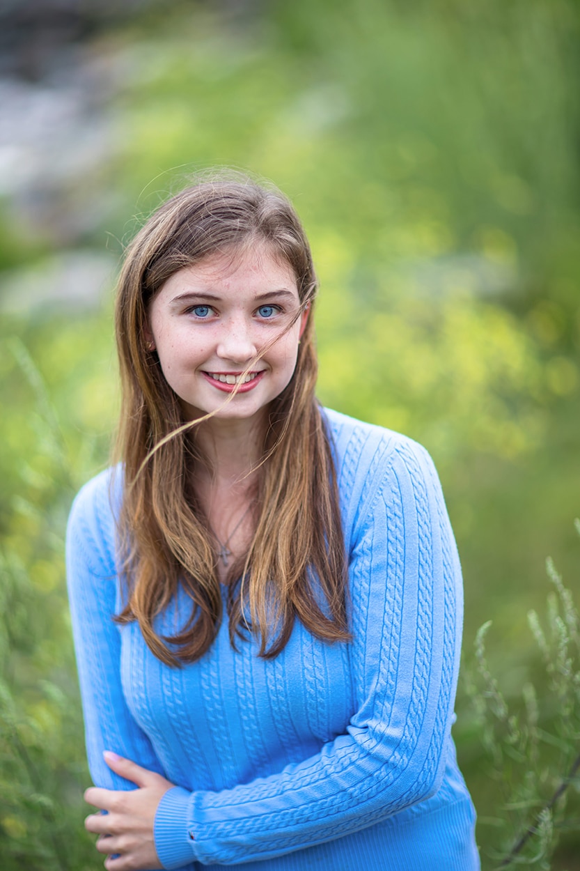 Senior portrait of a young woman in a blue sweater standing among wildflowers, playfully holding a blade of grass
