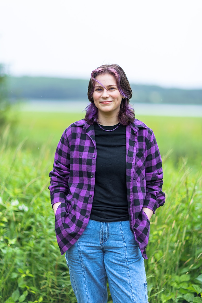 Senior portrait of a young woman in a purple plaid shirt and jeans standing in tall grass