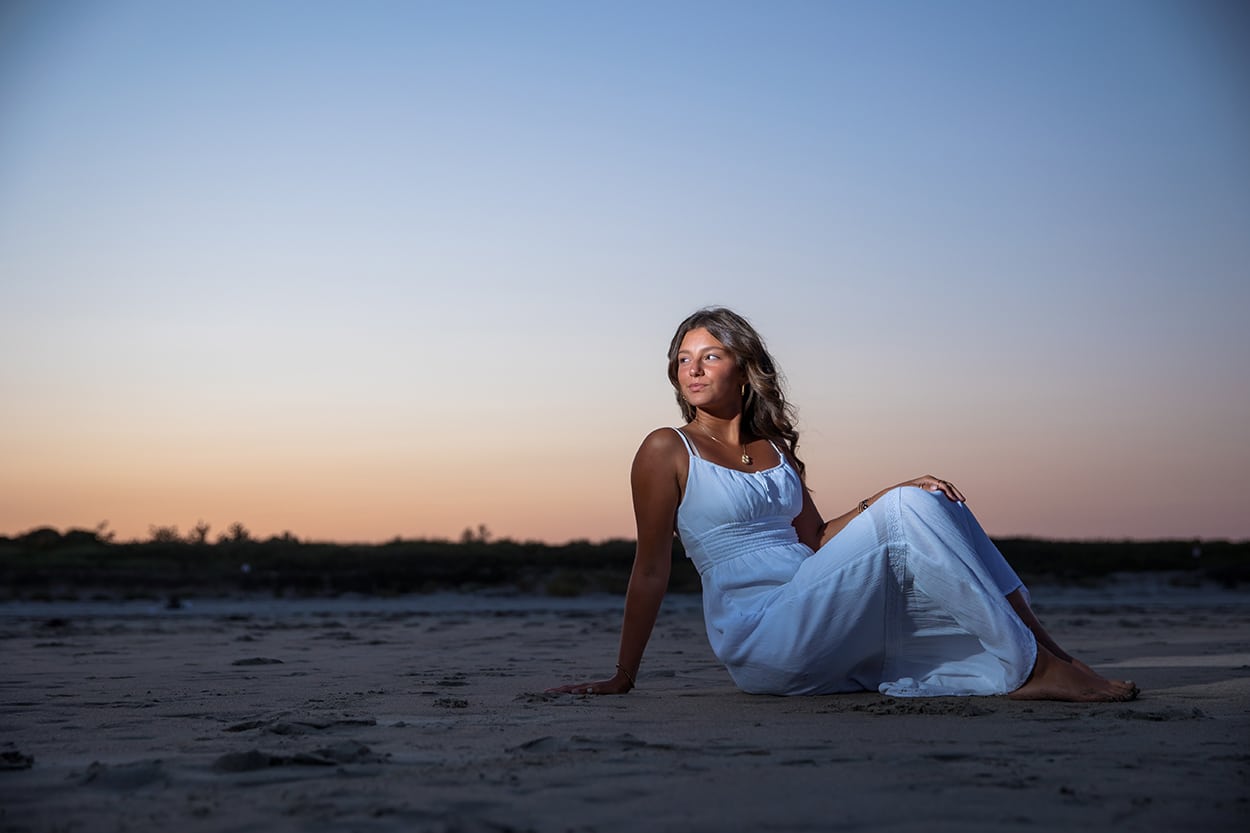 Senior portrait of a young woman in a flowing dress sitting on a beach at sunset