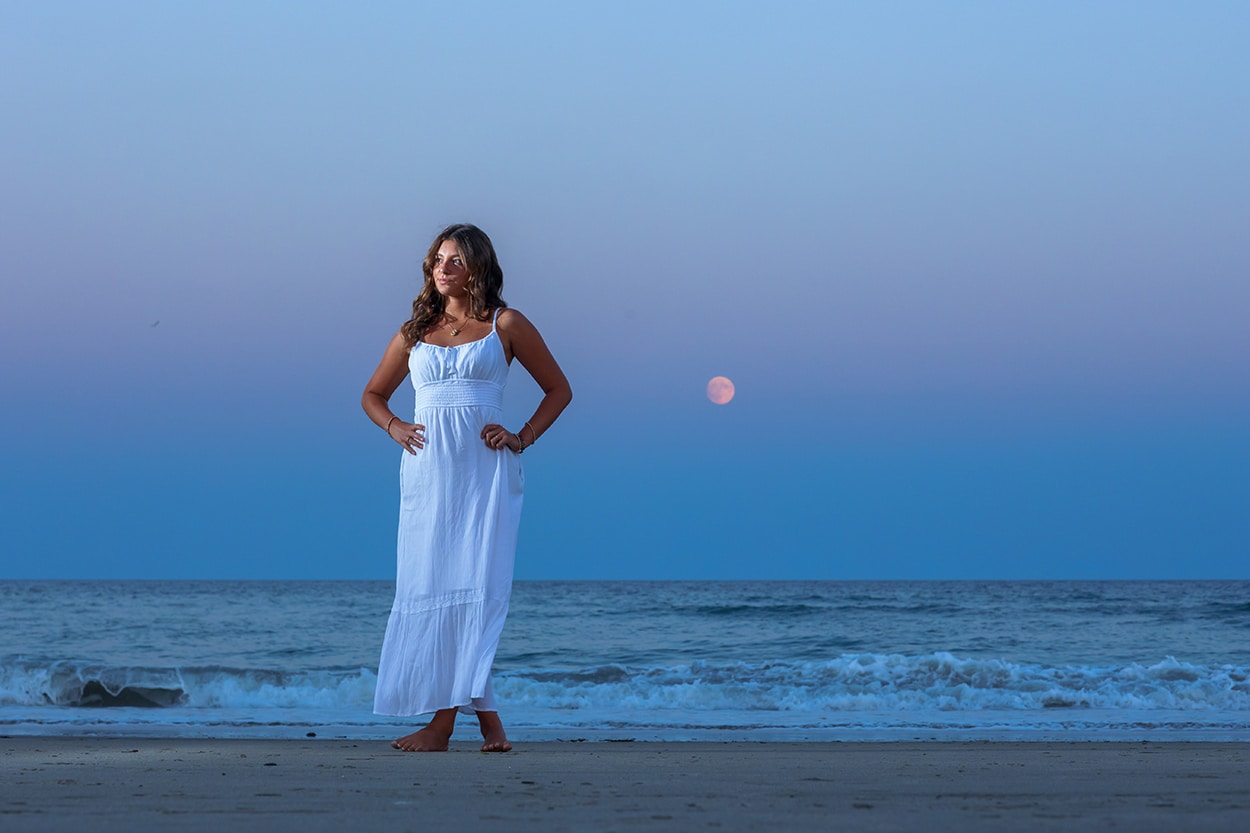 Senior portrait of a young woman in a white dress standing on a beach at dusk with the moon visible