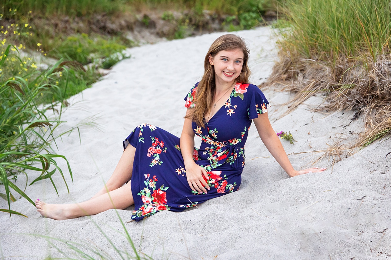 Senior portrait of a young woman in a floral dress sitting in the sand among dune grass