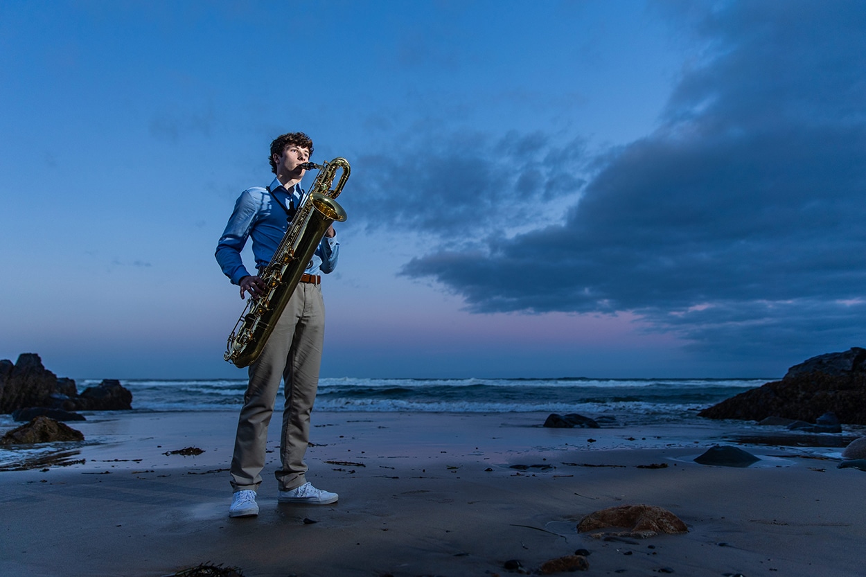 Senior portrait of a young man playing saxophone on a rocky beach at dusk