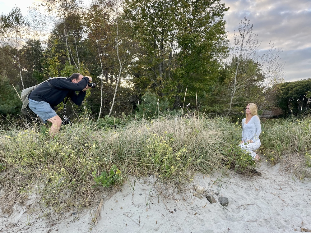 Behind-the-scenes photo of a photographer crouching on a grassy dune to photograph a young girl standing nearby